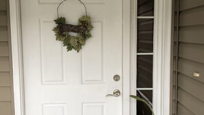 A white door with a wreath hanging on the doorknob, a potted plant beside it, and a black doormat in front.