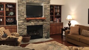 A living room with a stone fireplace, a television mounted above it, and a ceiling fan. There are two couches and a bookshelf filled with books.