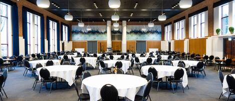 A large conference room with multiple round tables and chairs, high ceilings with hanging lights, and large windows along one wall.