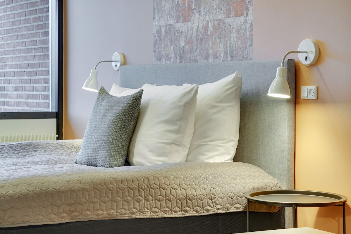 A neatly made bed with a quilted cover, two wall-mounted lamps, and a small round table beside it.
