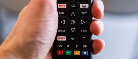 A hand holding a remote control with various buttons and icons, including Netflix and Prime Video.