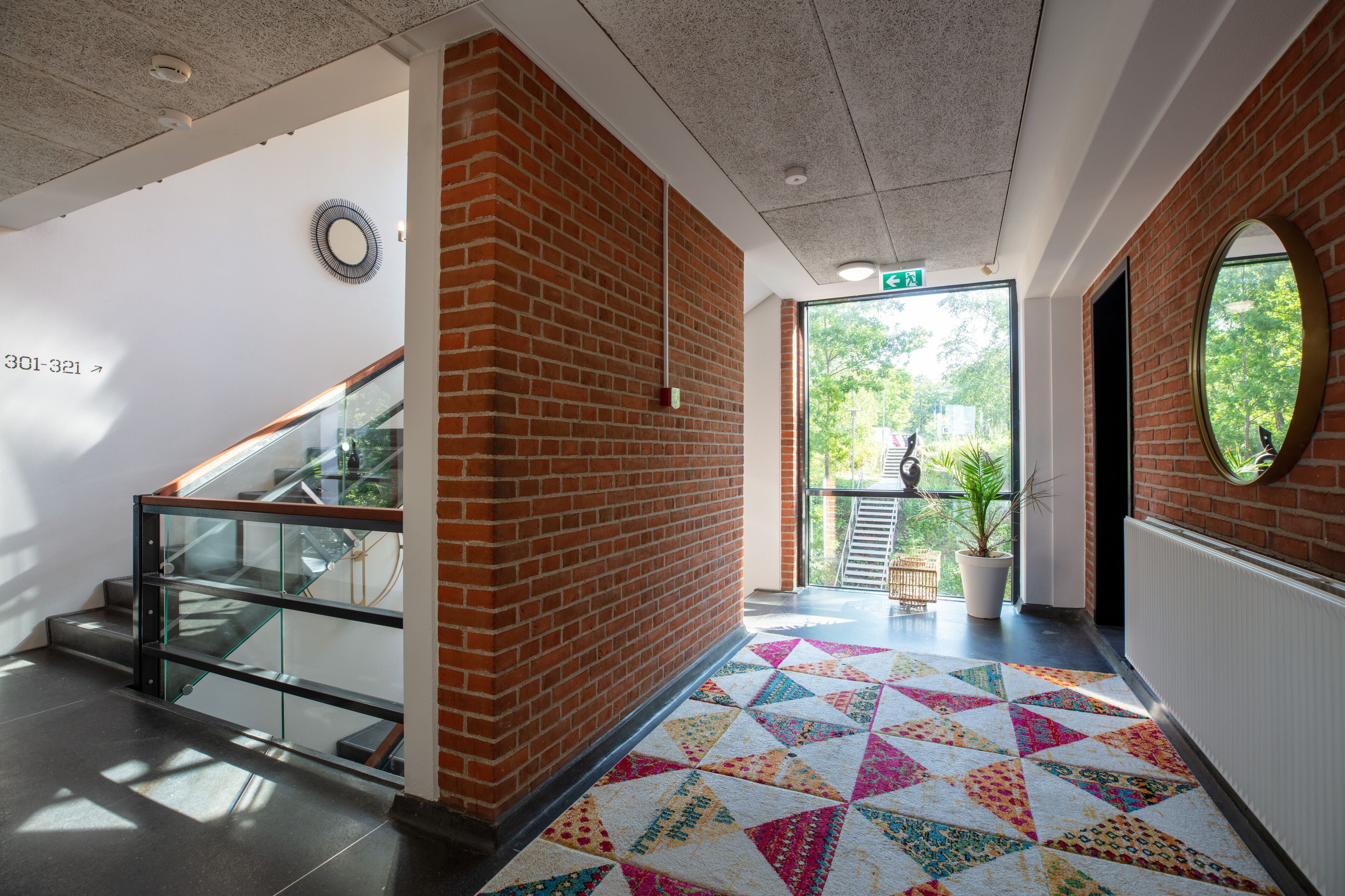 A modern interior with a brick wall, a glass staircase, a colorful geometric carpet, and a view of greenery outside.