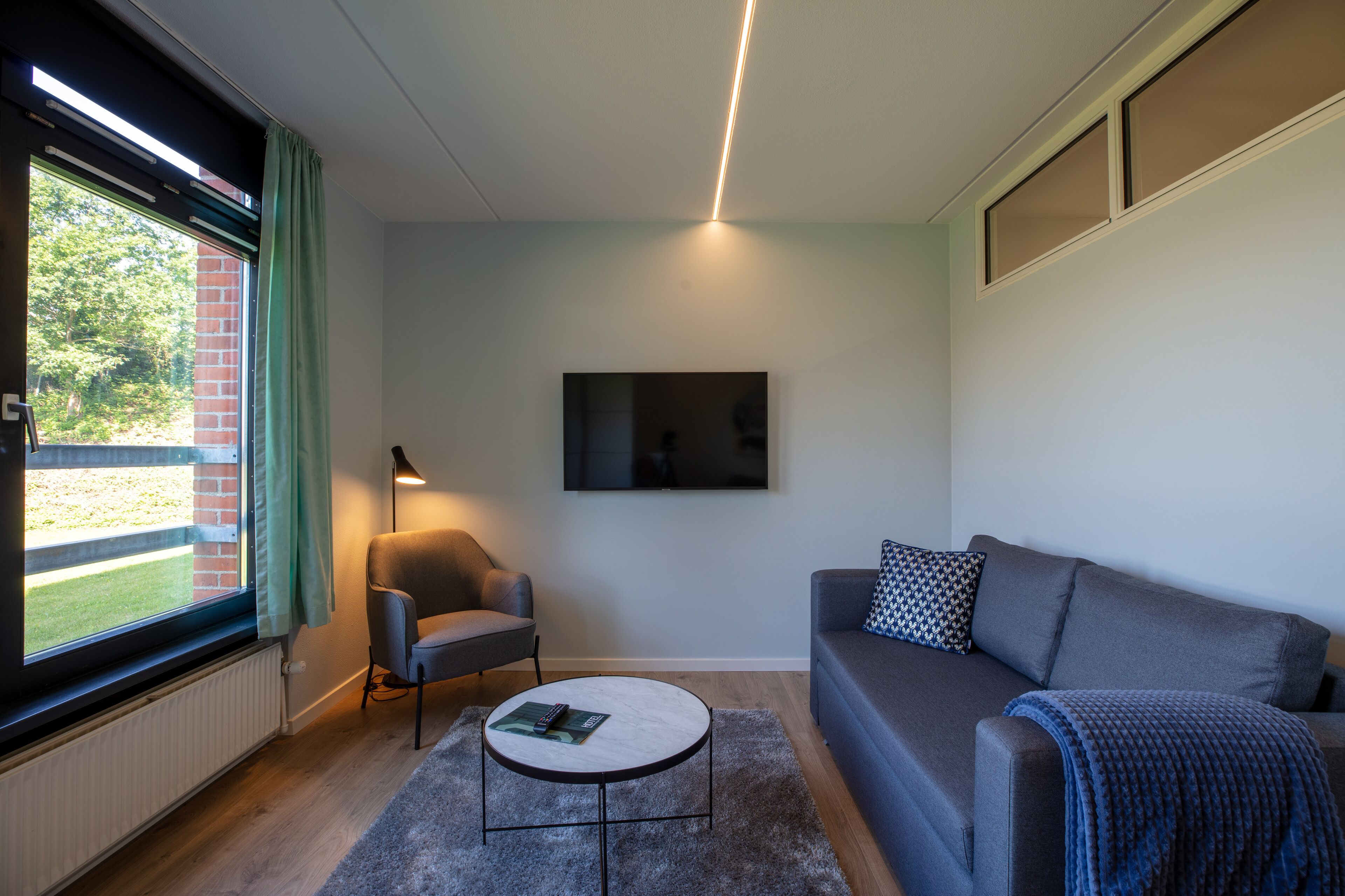 A modern living room with a grey sofa, a round coffee table, and a single armchair. A flat-screen TV is mounted on the wall, and a window with a view of greenery is visible.