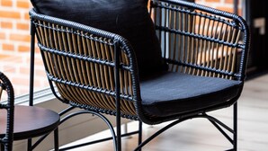 Una silla de mimbre con cojín negro en una habitación con pared de ladrillo y ventanas amplias.