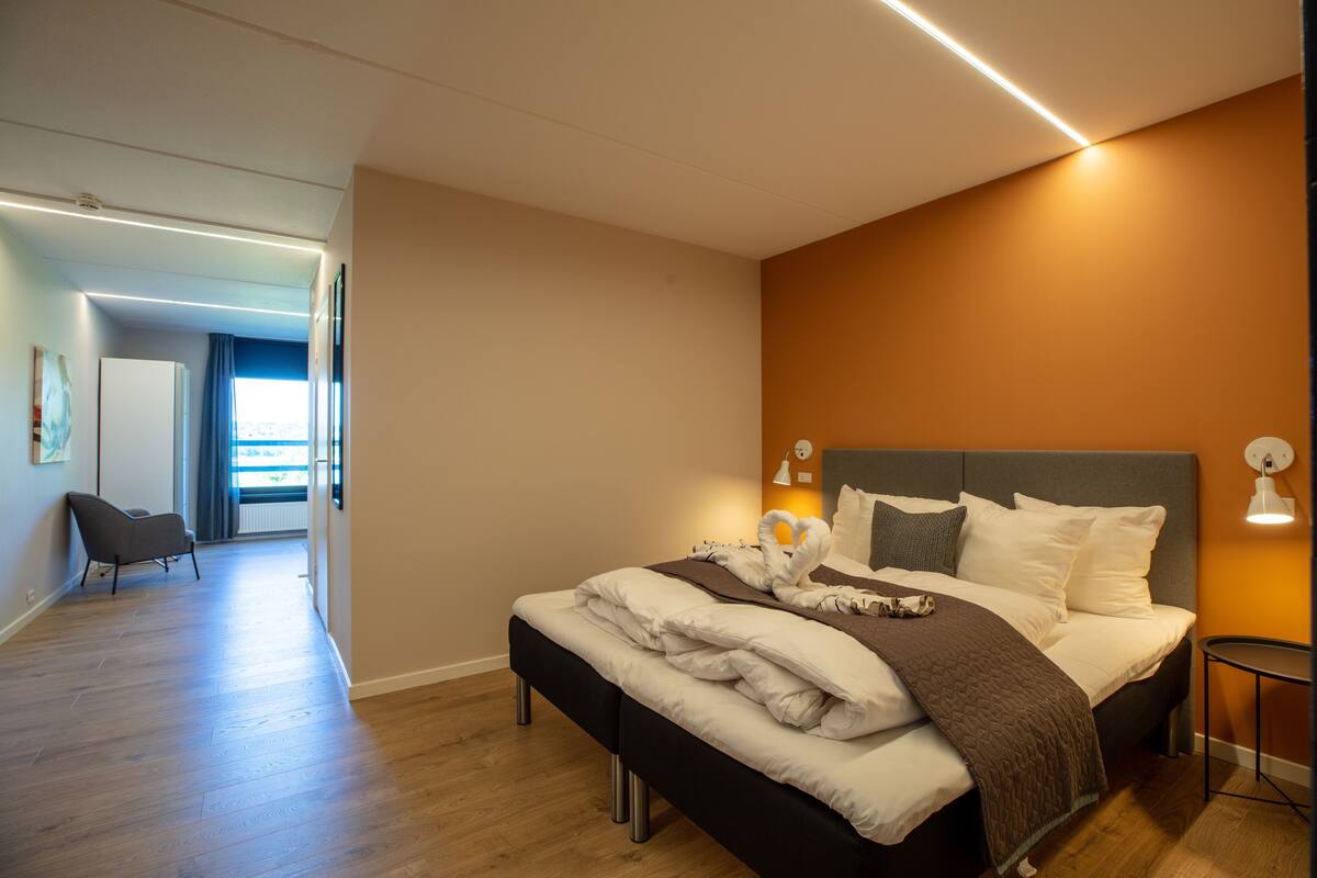 A modern hotel room with a large bed, a bedside table, and a view of the city through the window.