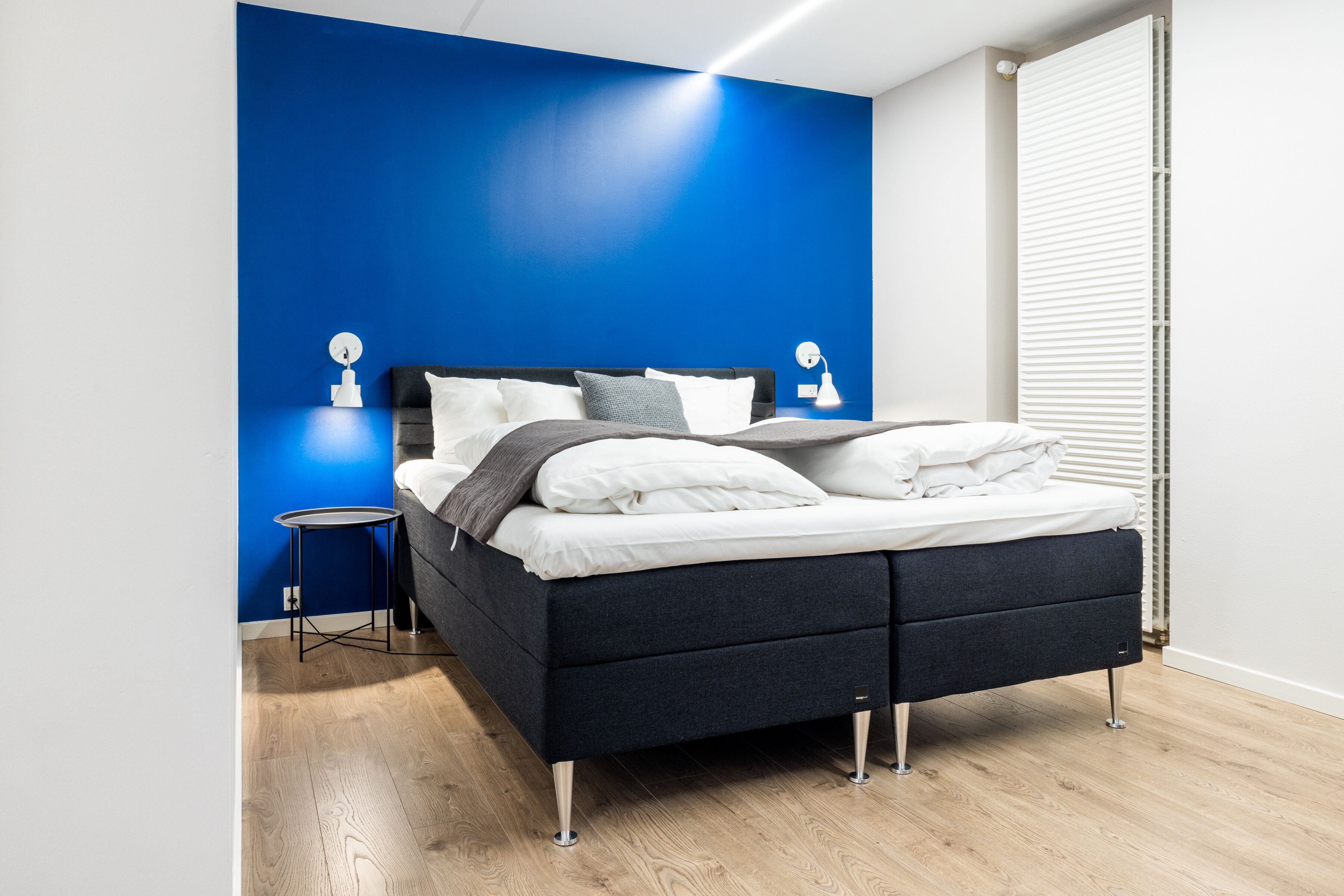A modern bedroom with a dark grey bed, white bedding, and a blue accent wall.