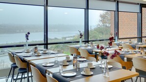 Un área de comedor con mesas dispuestas para el servicio, amplias ventanas que ofrecen vistas a un cuerpo de agua, y una pared de ladrillo al fondo.