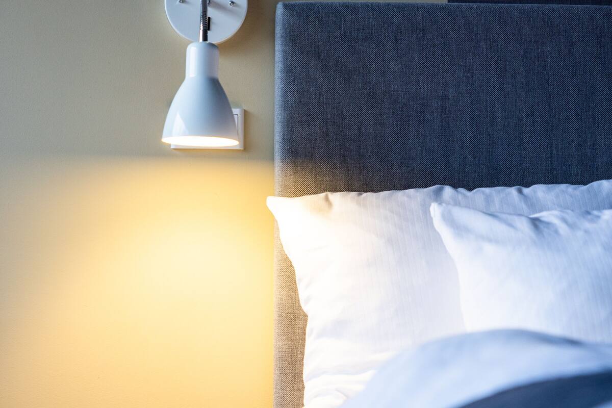 A neatly made bed with white linens, a round bedside table, and a wall-mounted lamp.