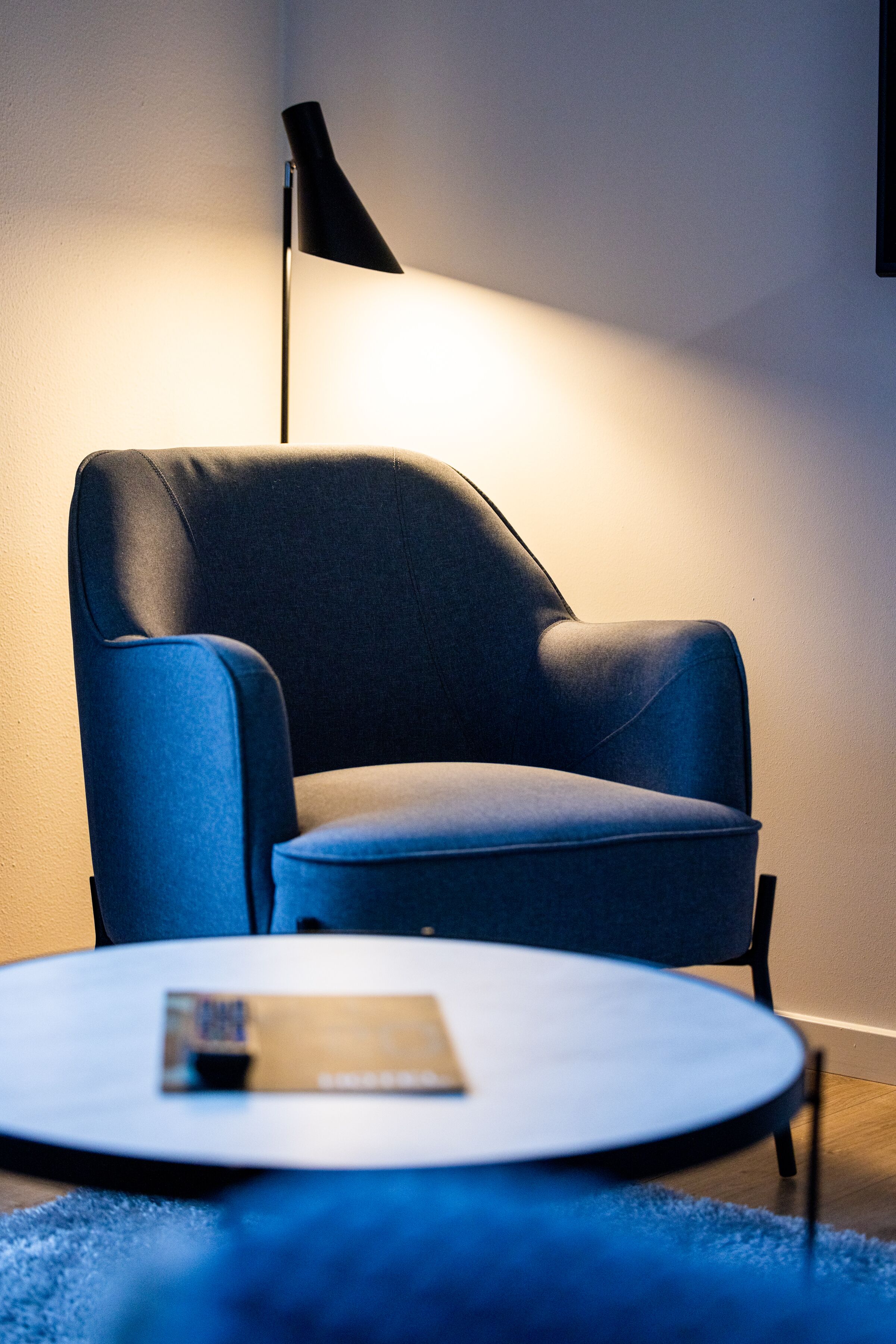 A modern room with a dark blue armchair, a round table with a remote control, and a black floor lamp.
