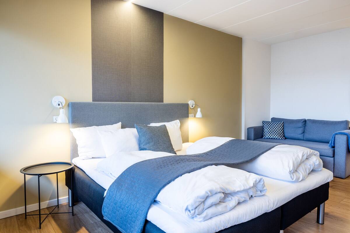 A modern hotel room with a large bed, a small side table, a sofa, and a wall-mounted lamp.