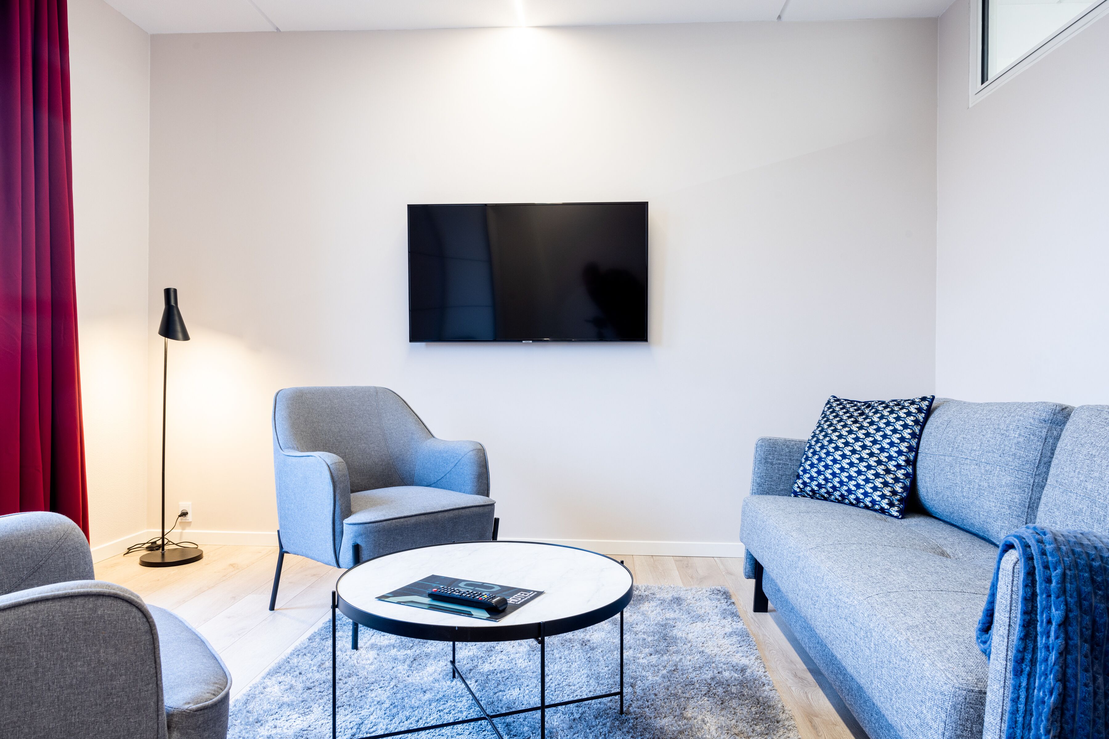 A modern living room with a flat-screen TV mounted on the wall, a grey sofa with patterned cushions, a blue armchair, and a round coffee table with a remote control.