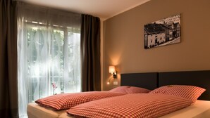 Een tweepersoonsbed met geruit beddengoed, een nachtkastje met lamp, een raam met dunne gordijnen, en een aan de muur bevestigd schilderij.