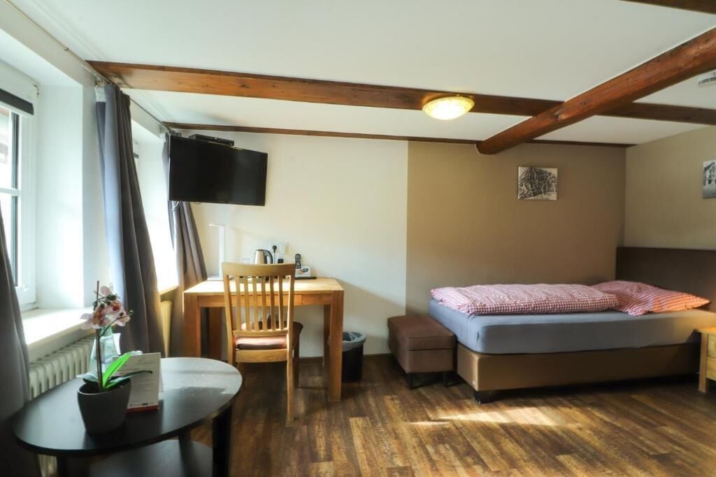 Ein kleiner Raum mit einem Bett, einem Schreibtisch, einem Sessel, einer Zimmerpflanze und einem Fernseher.