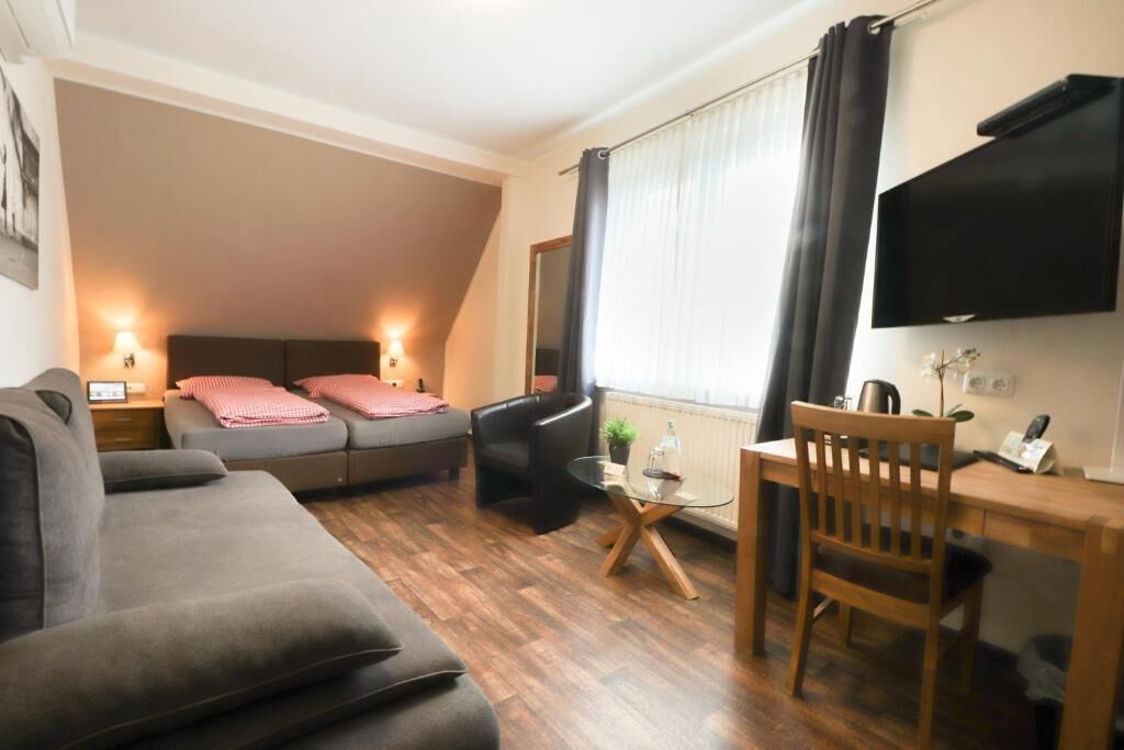 A modern room with a sofa, a bed with a headboard, a small table, a chair, a TV mounted on the wall, and a window with curtains.