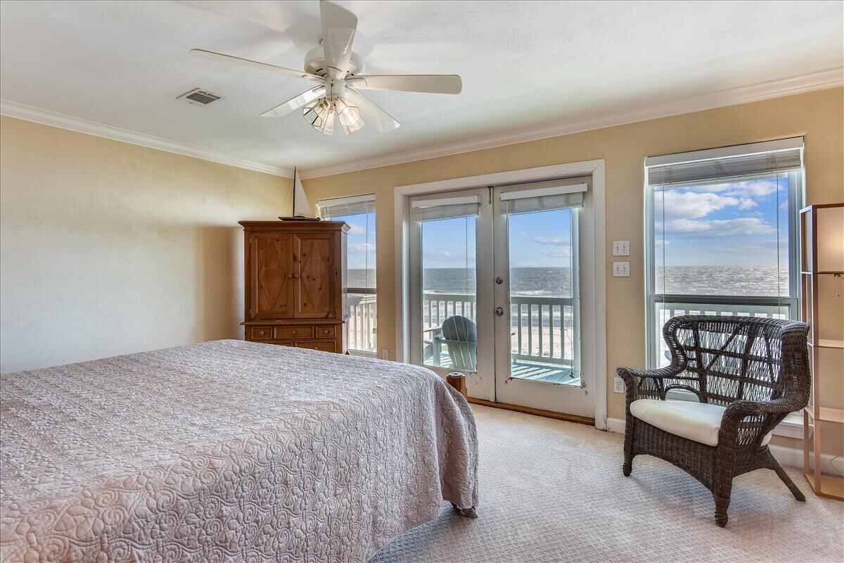A bedroom with a large bed, a ceiling fan, a wooden cabinet, a wicker chair, and a balcony with a view of the ocean.