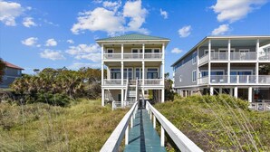 A wooden boardwalk leading to a beachfront property with two-story buildings, a covered walkway, and a view of the ocean. - LET AN IRISH SEABREEZE WISK YOUR CARES AWAY! (St. George Island)