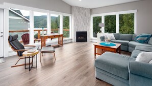 A spacious living room with a fireplace, a large sofa, a dining area with chairs, and a view of the outdoors through large windows. - Beautiful newer home in Sudden Valley offering stunning lake view, golf and more (Bellingham)