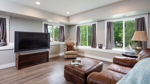 A modern living room with a flat-screen TV, leather furniture, and large windows offering a view of greenery. - Beautiful newer home in Sudden Valley offering stunning lake view, golf and more (Bellingham)