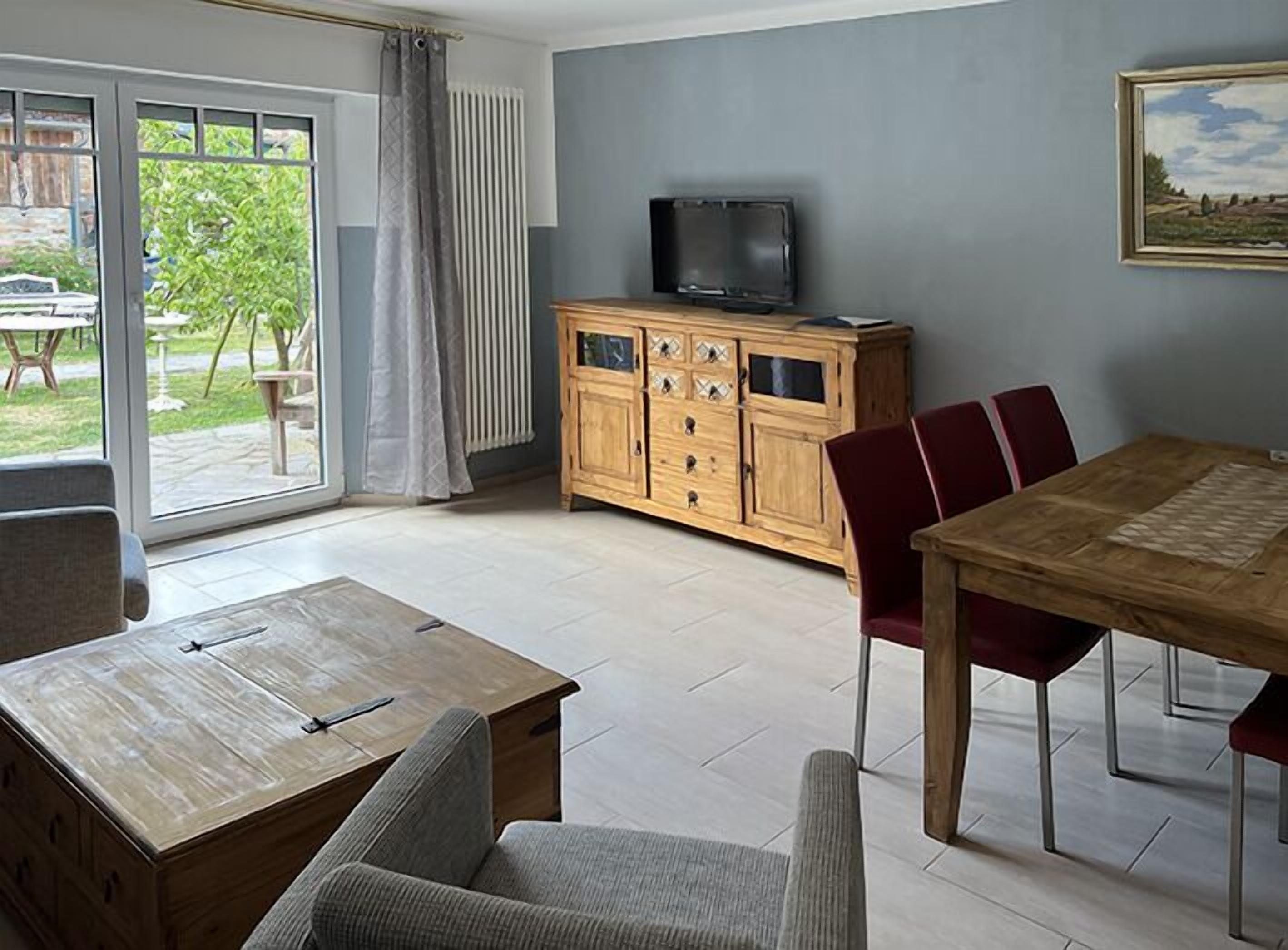 A modern living room with a wooden dining table, red chairs, a TV on a wooden cabinet, and a sliding glass door leading to a patio.
