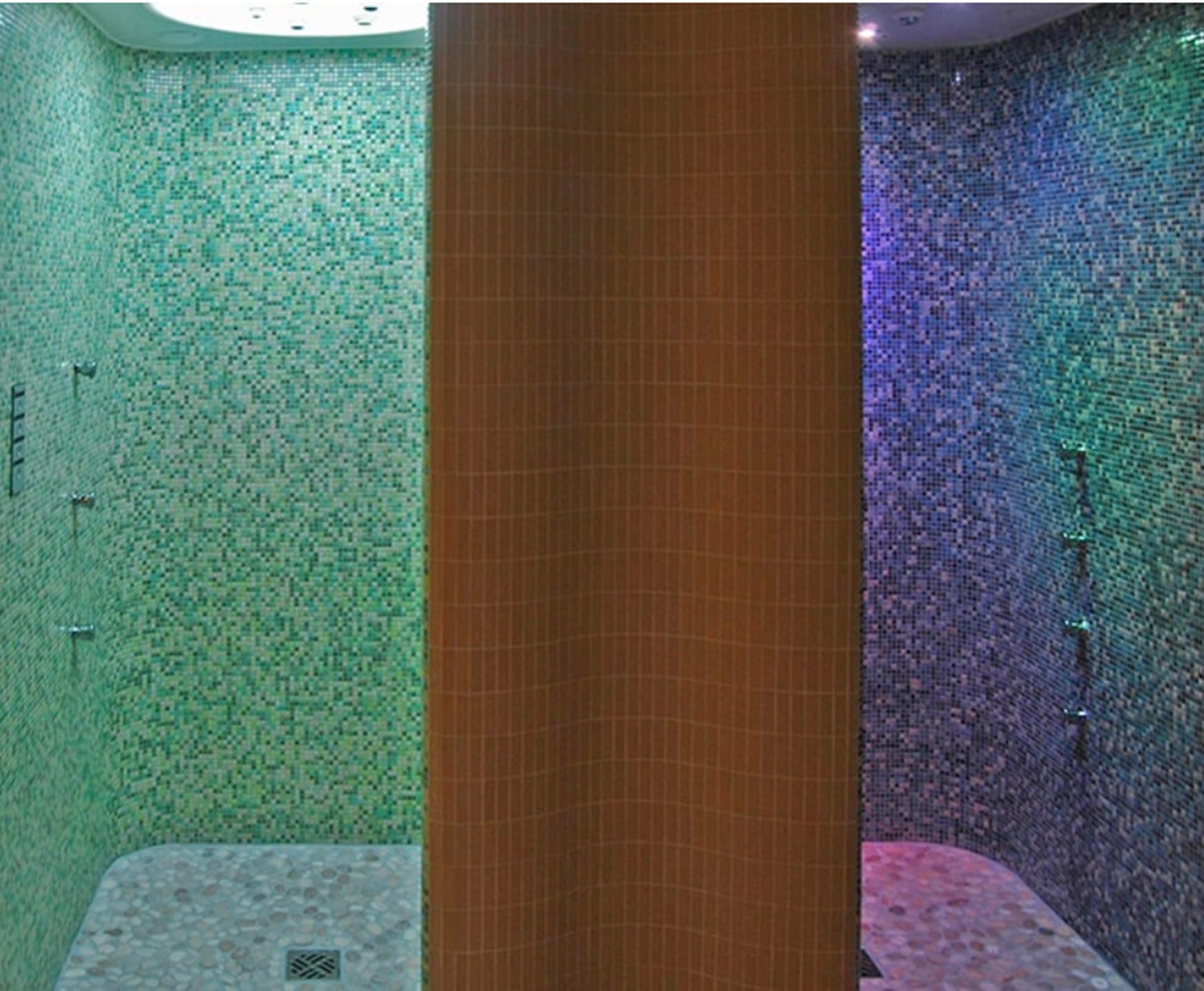 Une salle de bain aux murs recouverts de mosaïques vertes et bleues, avec une colonne en bois et un espace douche doté d’un sol composé de petits galets.