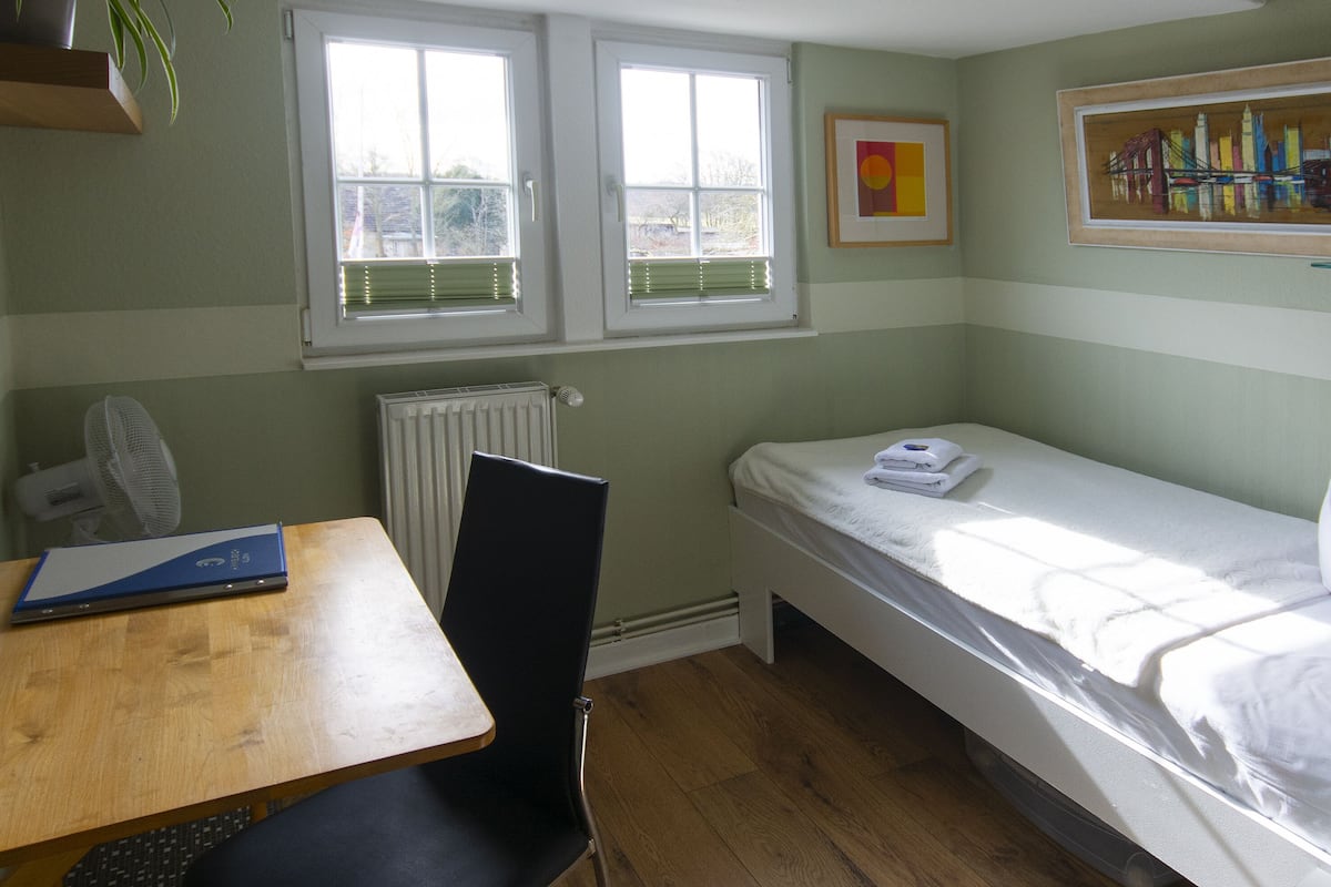 A small, clean room with a single bed, a desk, and a chair. There are two windows with blinds, a radiator, and a framed picture on the wall.