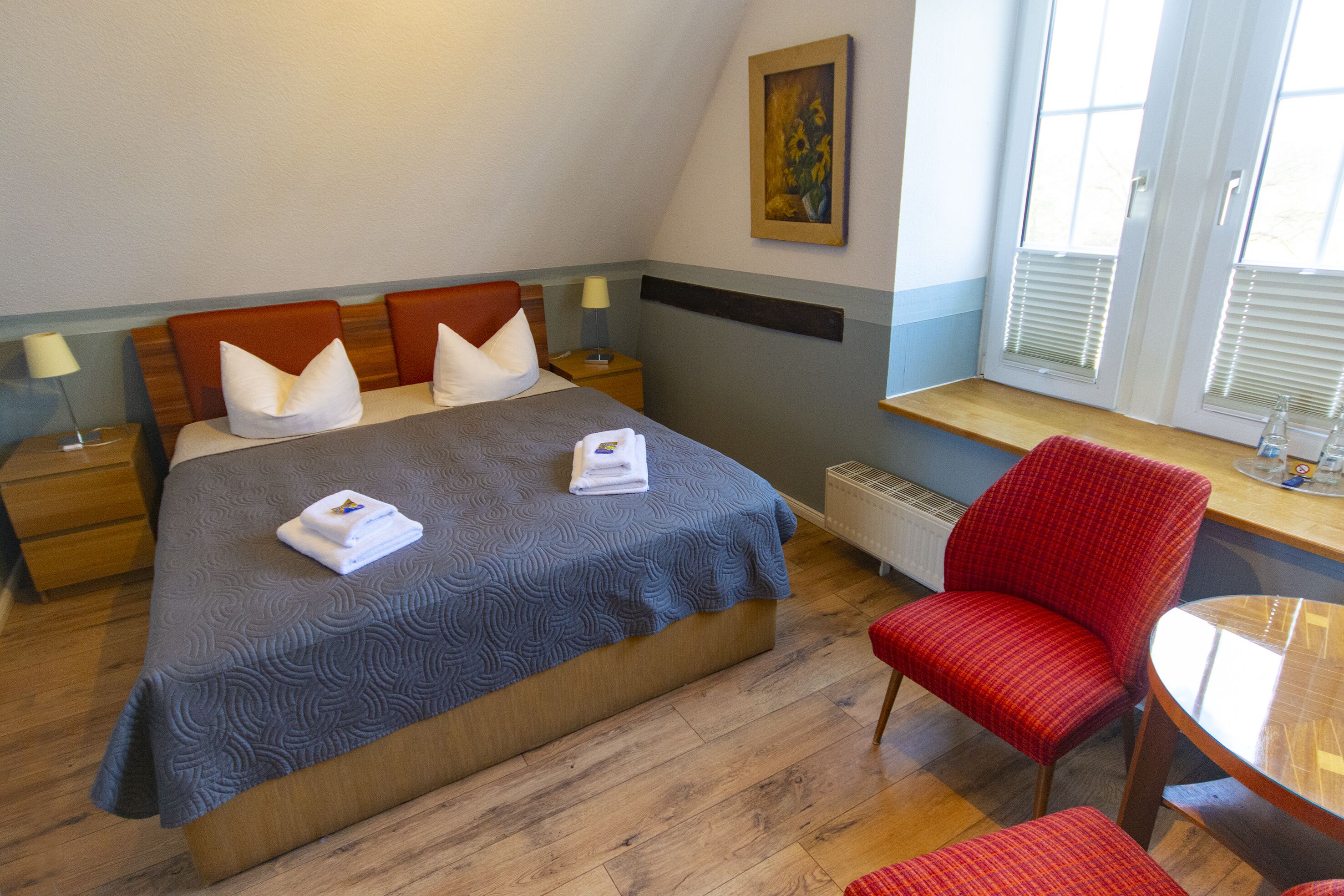 A hotel room with a bed, two nightstands, a red chair, a small table, and a window with blinds.