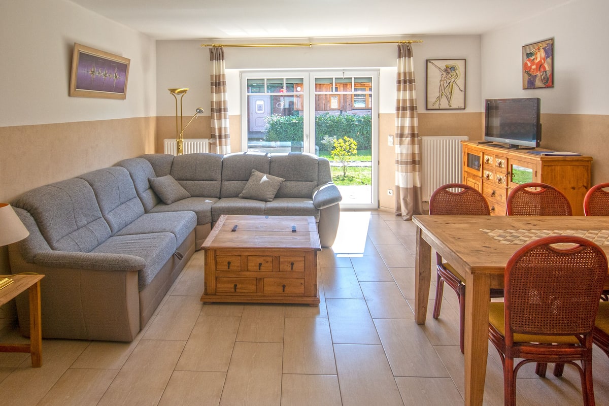 A living room with a gray sectional sofa, a wooden coffee table, a dining table with chairs, a television on a wooden cabinet, and a view of a garden through a glass door.