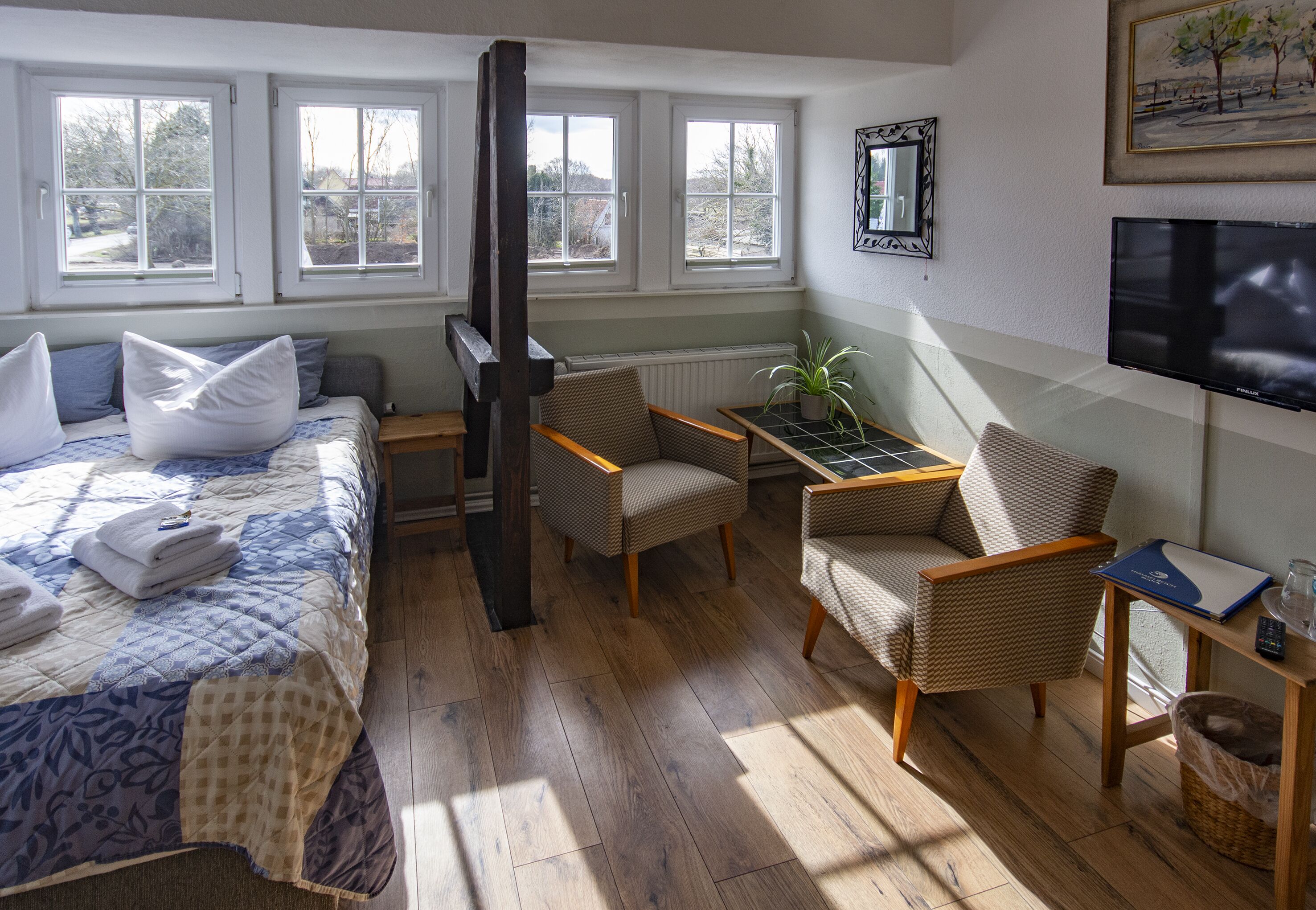 Ein Zimmer mit einem Bett, zwei Sesseln, einem kleinen Tisch, einem Fernseher und einem Fenster mit Blick auf Bäume.