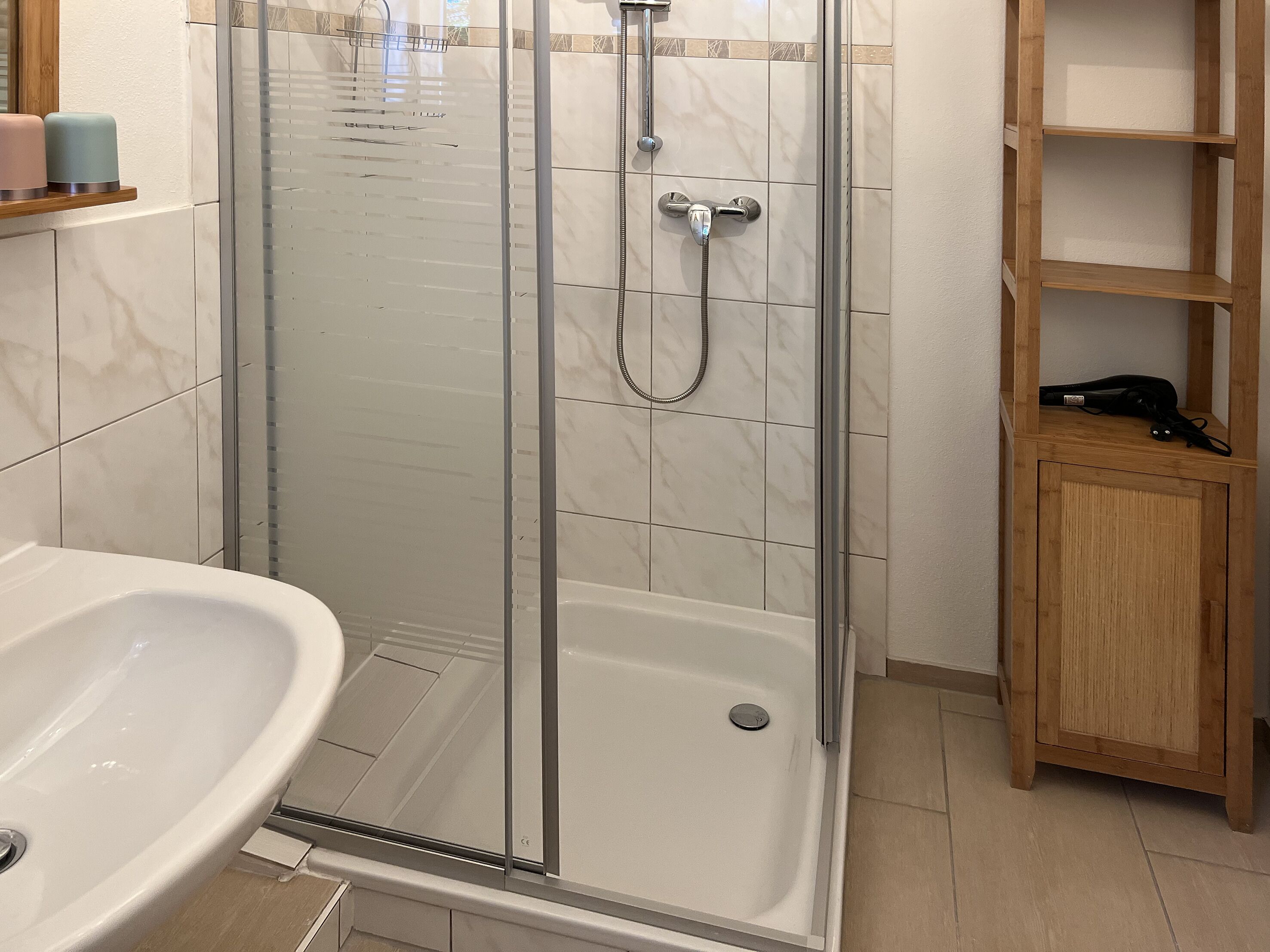 Une salle de bain avec une douche à parois de verre, un lavabo blanc et une étagère en bois où sont rangés les articles de toilette.