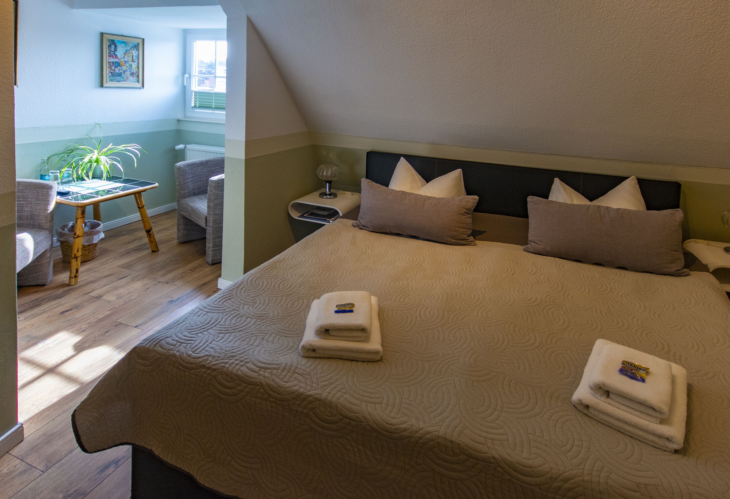 A neatly made bed with two folded towels, a bedside table with a lamp, and a chair in a room with wooden flooring and a window.