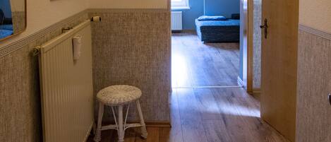 Een gang met houten vloer, een rieten krukje en een radiator. Er hangen prenten in de muren en een deur die naar een andere kamer leidt.
