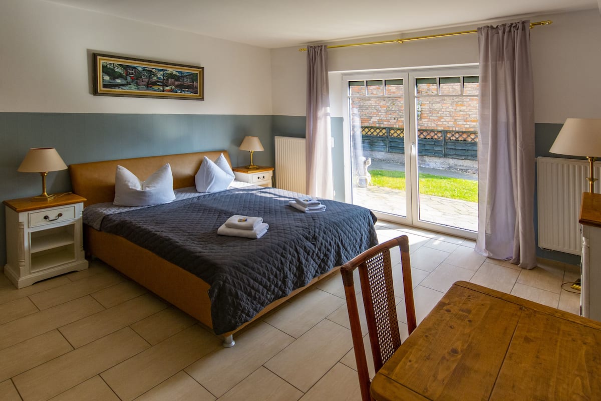 A hotel room with a bed, two bedside tables with lamps, a wooden chair, a wooden table, and a view of a garden through a sliding glass door.