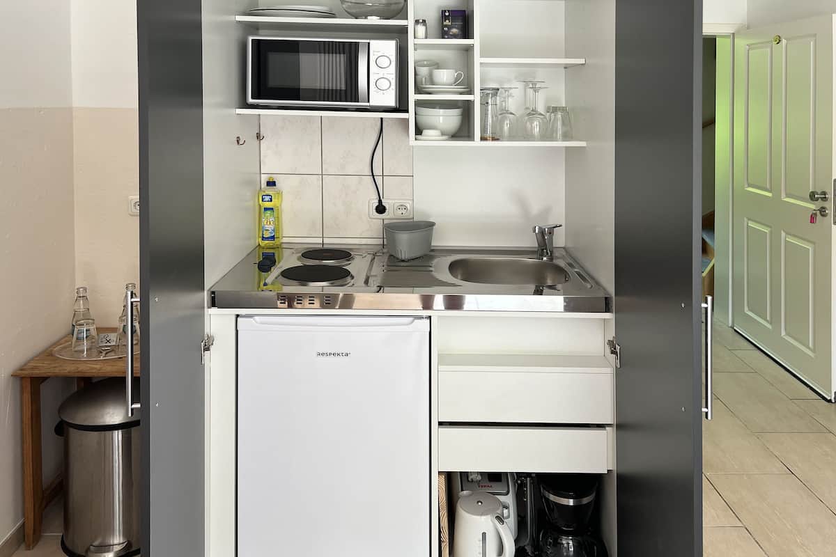 A compact kitchen with a microwave, sink, and coffee maker, featuring white cabinetry and a small trash can.