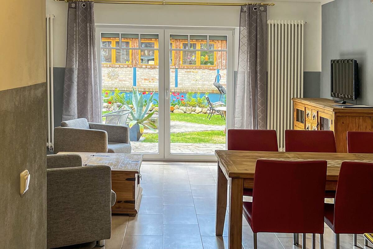 A modern living room with a sliding glass door leading to a patio, a wooden dining table with red chairs, and a television on a wooden cabinet.