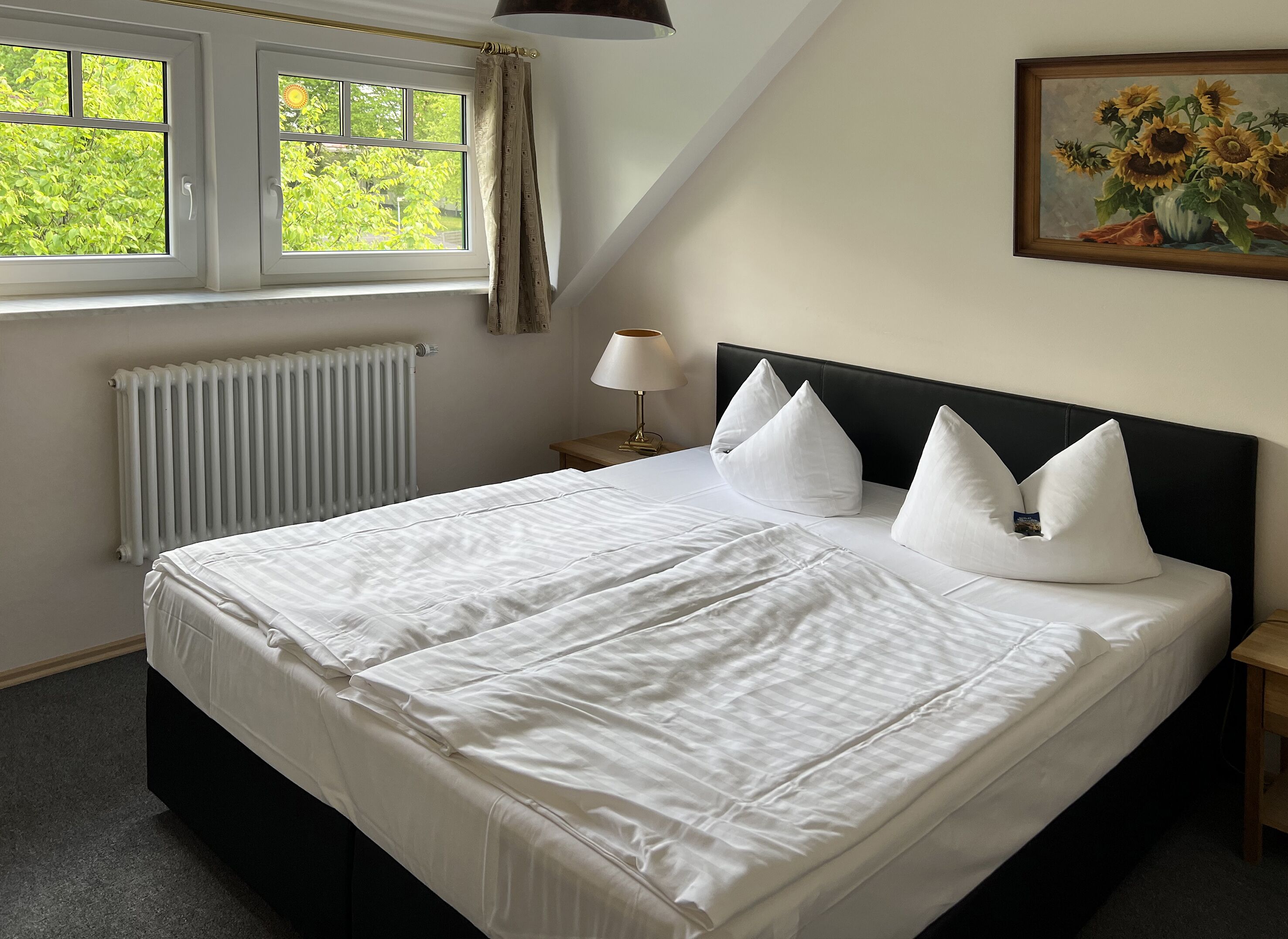 Un lit bien fait, avec du linge de lit blanc, une tête de lit noire et trois oreillers blancs. Il y a une fenêtre avec des rideaux, un radiateur et un tableau représentant des tournesols accroché au mur.