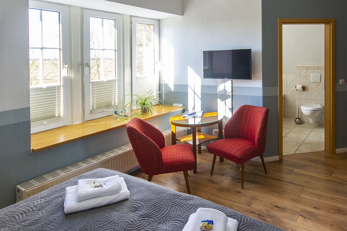 A hotel room with a bed, two red chairs, a small table, a TV, and a window with blinds.