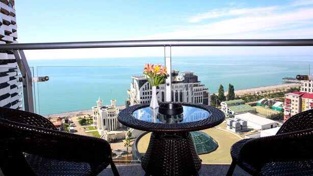 Un balcón con sillones de mimbre y una mesa con superficie de vidrio, con vista a la playa y al paisaje urbano.