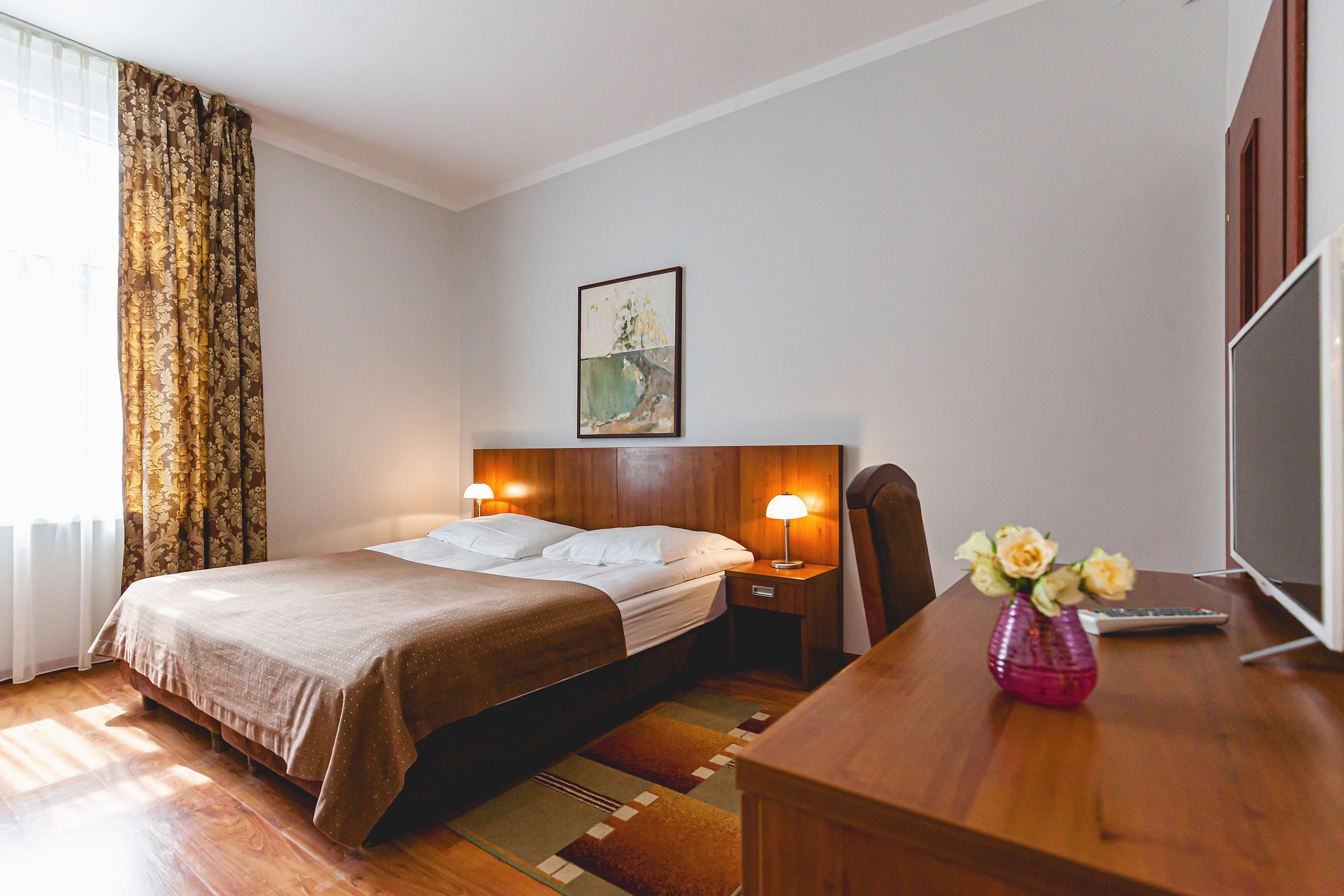 A hotel room with a bed, bedside lamps, a desk with a vase of flowers, a television, and a painting on the wall.