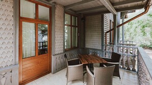 Un espace extérieur abrité, avec une table en bois et des chaises en osier, une porte en bois et un balcon doté d’une balustrade.