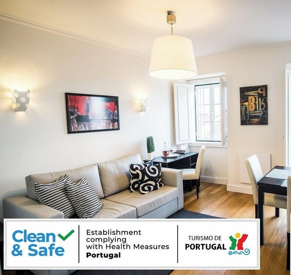 A modern living room with a sofa, dining table, and artwork. There are signs indicating health safety measures and a QR code for more information.