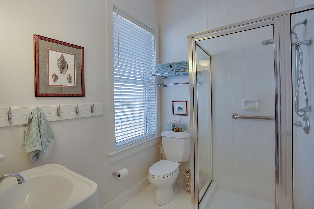 A bathroom with a white sink, toilet, and glass-enclosed shower. There is a window with blinds, a framed picture on the wall, and a towel rack.