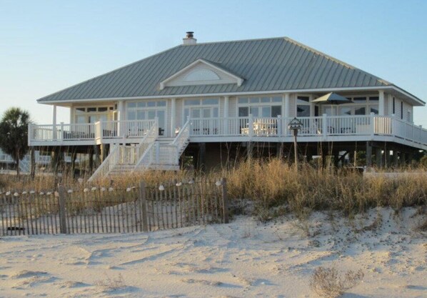 A beachfront house with a covered porch, a staircase leading to the upper level, and a sandy beach in the foreground.