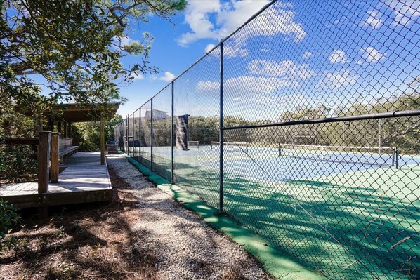 A fenced tennis court with a wooden deck and a covered seating area.