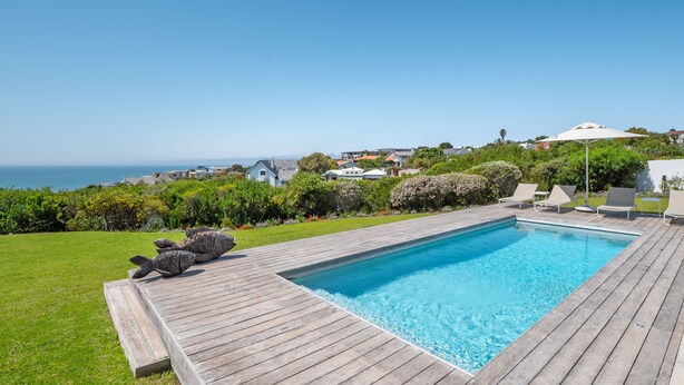 Una piscina rectangular con un muelle de madera, rodeada de césped y vistas al océano.