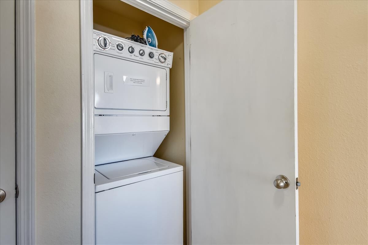 Une petite buanderie équipée d’un lave-linge et d’un sèche-linge superposés, d’une petite planche à repasser et d’une porte dotée d’une poignée argentée.