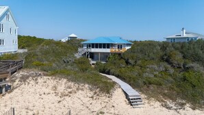 Una propiedad frente a la playa con una casa destacada, techumbre azul, muelle de madera y una escalera que lleva a un balcón.