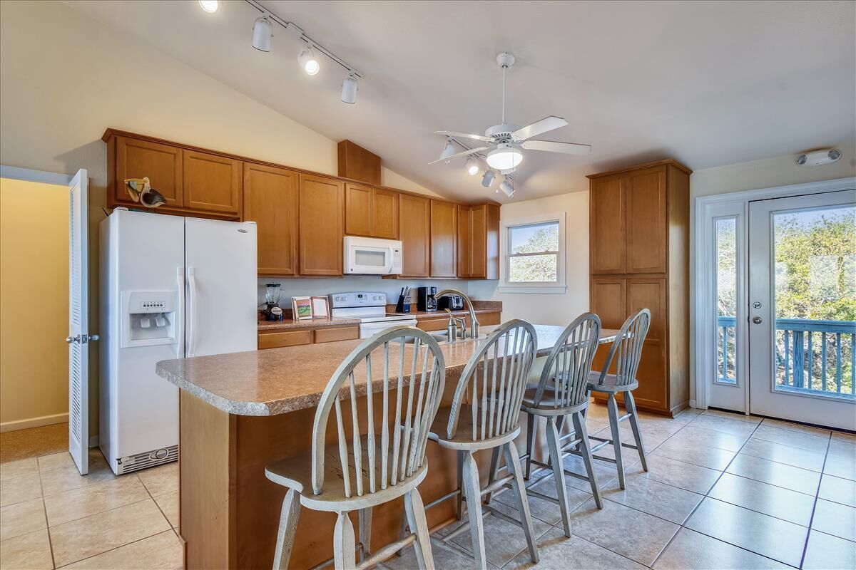 A kitchen with wooden cabinets, a refrigerator, a microwave, a ceiling fan, and a bar area with chairs.