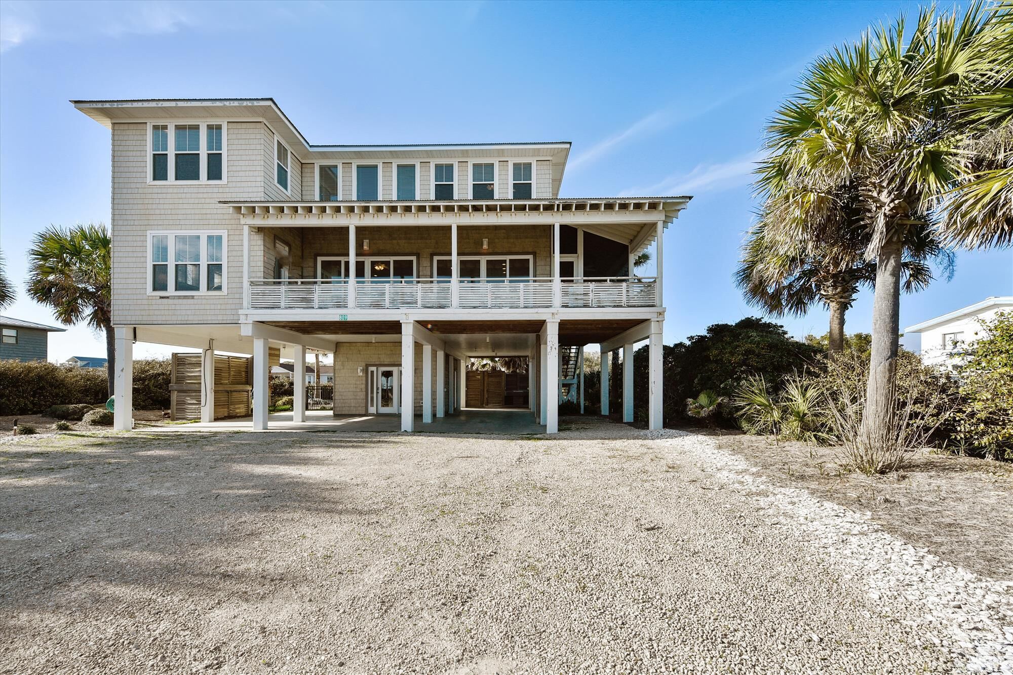 A two-story beachfront house with a balcony, a covered entryway, and a gravel driveway.