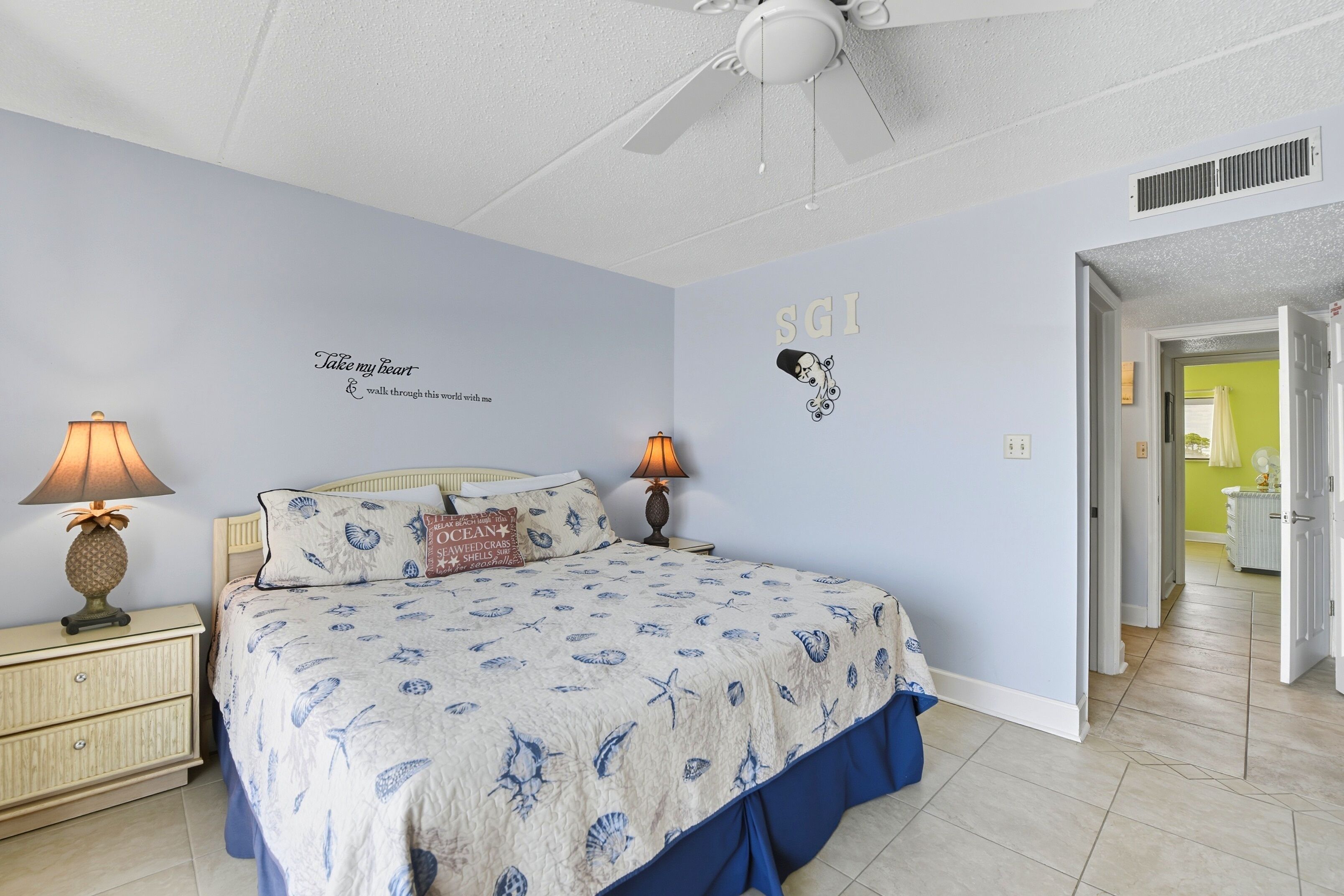A bedroom with a bed, two nightstands with lamps, a ceiling fan, and a wall with a marine-themed bedspread and decorative items.
