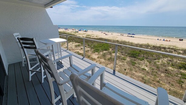 A balcony with white chairs and a table, overlooking a beach with people and umbrellas.